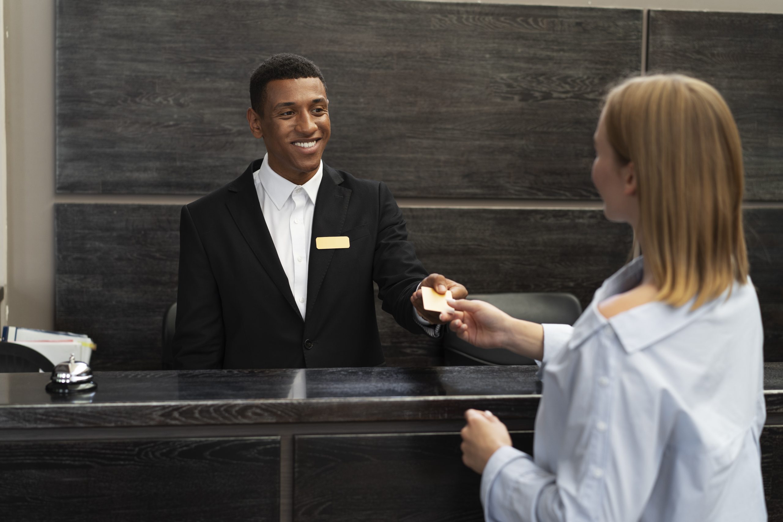 Hotel receptionist smiling, handing room keycard to a guest at the front desk.